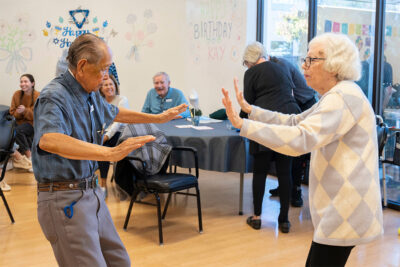 Two elderly people are dancing together in a room decorated with colorful "Happy Birthday" signs. Other people in the background are seated and smiling.